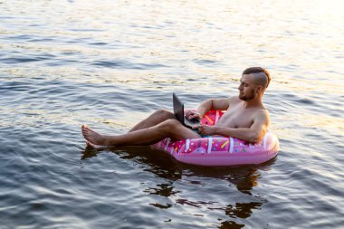 Şişme bir halka üzerinde otururken bir dizüstü bilgisayar üzerinde çalışan işkolik adam, kopya alanı.