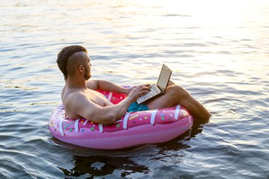 Şişme bir halka üzerinde otururken bir dizüstü bilgisayar üzerinde çalışan işkolik adam, kopya alanı.