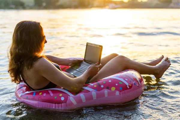 Mujer de negocios freelancer trabajando en un portátil mientras está sentado en una dona inflable de goma, sobre un fondo de puesta de sol . — Foto de Stock