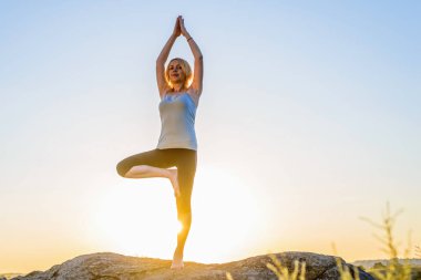 Gün batımında dağların tepesinde ağaç yogası pozisyonunda meditasyon alan kadın