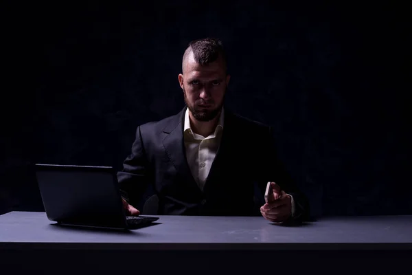 Серйозний бізнесмен, сидячи за столом, працює на ноутбуці і розмовляє на смартфоні, на темному тлі . — стокове фото