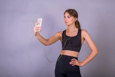 Gri arka plan üzerinde stüdyoda kulaklık ve akıllı telefon kullanarak müzik dinleyen genç kadının portresi.