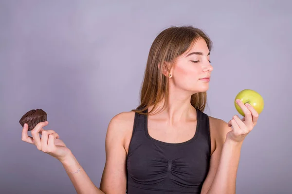 Right choice. Studio shot of young fitness woman in sports clothing holding green apple in one hand and cake in another.