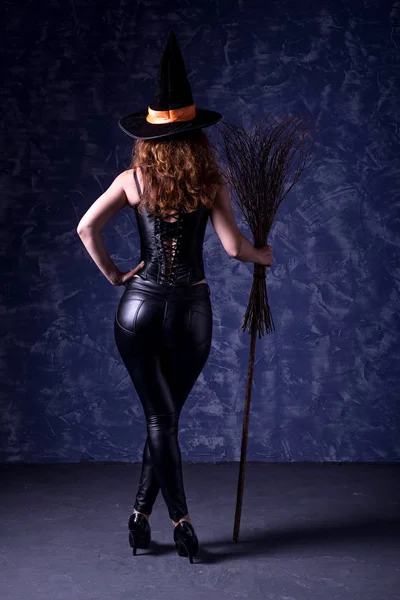 Deri pantolon ve koyu bir arka plan üzerinde poz bir süpürge ile bir korse seksi kadın, arka görünüm.