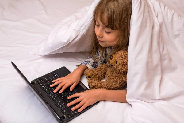 Linda niña usando un portátil en la cama debajo de una manta . — Foto de Stock