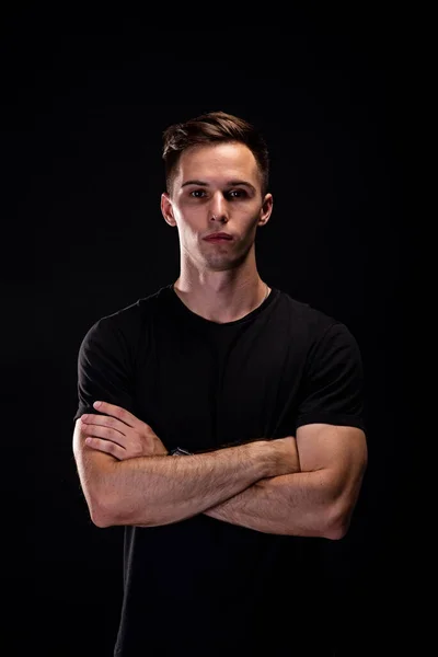 Portrait of a young athletic man in a black T-shirt on a black background. Young beautiful sporty man. Fitness lifestyle.