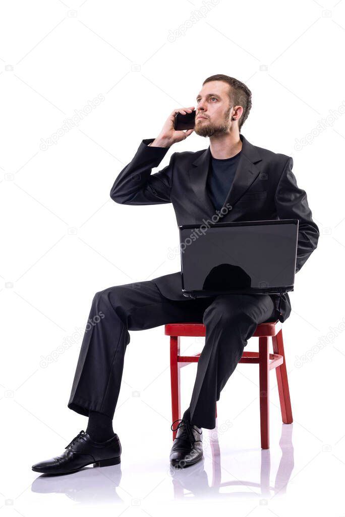 A bearded business man sits on a chair, holds a laptop and talks on the phone, on a white background. Business style.