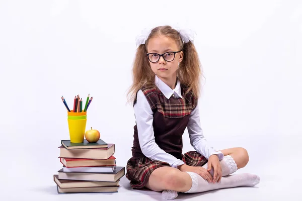 A little girl in a school uniform and glasses sits on a white background next to a pile of books. School concept.