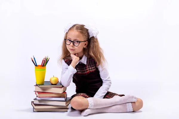 A little girl in a school uniform and glasses sits on a white background next to a pile of books. School concept.