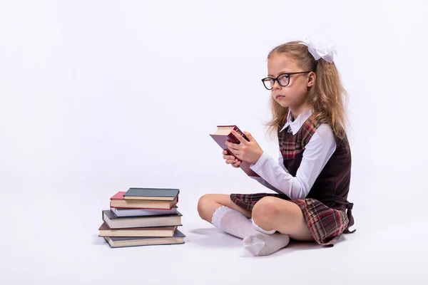 Little girl in school uniform chooses a book while sitting on a white background. School concept.