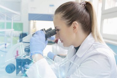 Genç bayan bilim adamı mikroskopla bir çalışmanın içine bakıyor.