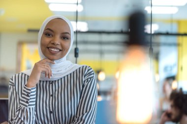 Başörtüsü takan Müslüman iş kadını ofiste bilgisayar başında çalışıyor. Başörtülü güzel Afrikalı kadın kameraya bakıyor ve gülümsüyor. İş yeri ve çok ırklı ilişkiler kavramı
