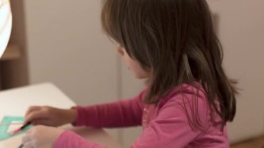 Okul masasında aydınlanmış bir küreyle resim çizen ve tablet izleyen sevimli küçük kız, seçici bir odak noktası.