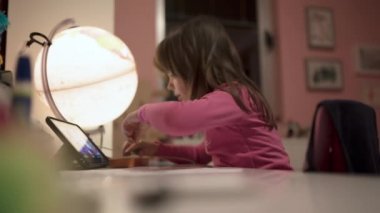 Okul masasında aydınlanmış bir küreyle resim çizen ve tablet izleyen sevimli küçük kız, seçici bir odak noktası.