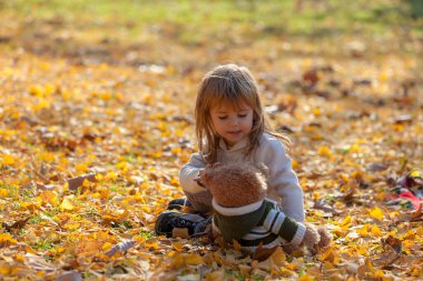 Küçük kız ayısıyla sonbahar yapraklarında oturuyor.