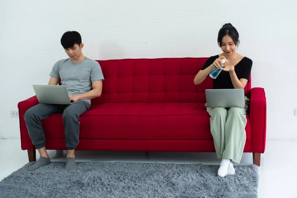 Happy Asian couple cleaning hands with sanitizer alcohol gel while sitting working together on sofa at home.