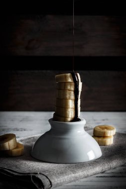 Lezzetli tatlı taze muz dilimleri sıvı dilimleri bir çul ve ahşap kurullarının bir arka plan üzerinde beyaz bir kabın üstüne çikolata ile ayakta. Dikey görüntü karanlık moody tarzı.