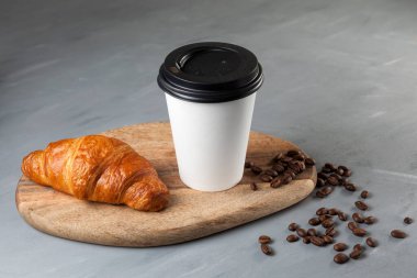 Taze kruvasan ve beyaz kağıt bardakta kahve gri arka planda tahta bir kesme tahtasının üzerinde. Yakınlarda birkaç kahve çekirdeği var..