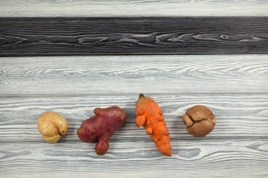 Multi-colored ugly vegetables (potatoes and carrots) lies on one row on a wooden textured background. Close-up, top view. Concept - reduction of food organic waste. clipart