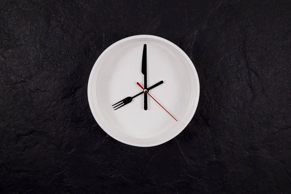 White plate clock on a dark textured stone background with copy space. The hands point to 8 o'clock. Concept - interval fasting or autophagy.