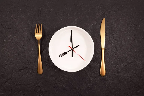 Clock hands on a white plate. Gold fork and knife on a dark stone background. Concept of autophagy or interval fasting.