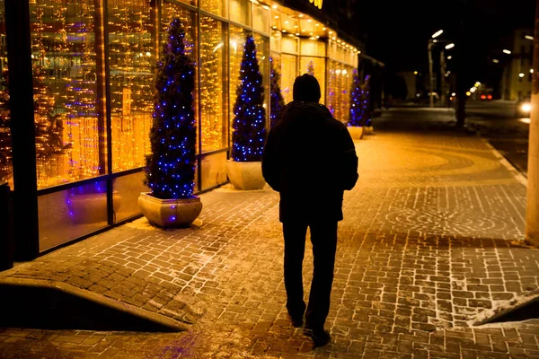 Man back to the viewer is on the night street, decorated with lights for Christmas.