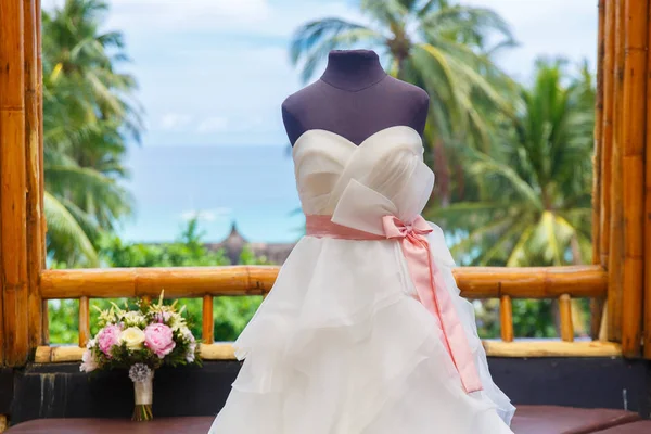 Wedding accessories. Mannequin in a wedding dress, and a bouquet of the bride on the window of a bamboo hut. Tropical landscape in the background, palm trees and the sea.
