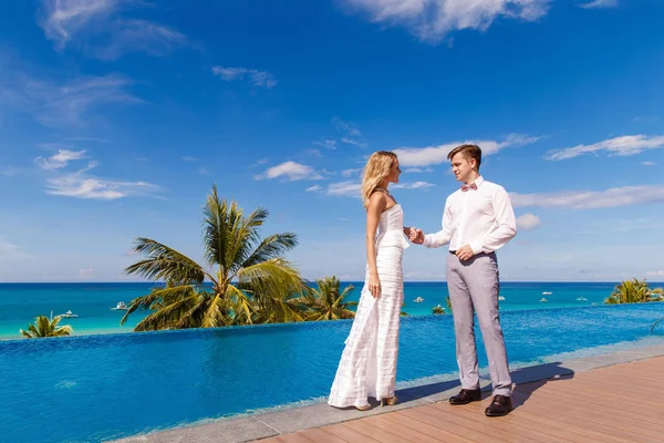 穿着白色婚纱的美丽金发新娘和新郎跳舞在泳池附近的旅馆 热带海洋 天空和棕榈树在背景中 夏季度假概念 — 图库照片