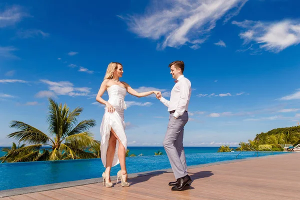 穿着白色婚纱的美丽金发新娘和新郎跳舞在泳池附近的旅馆 热带海洋 天空和棕榈树在背景中 夏季度假概念 — 图库照片