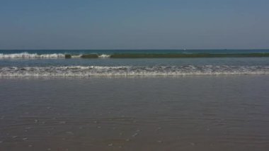 Sakin denizden dalgalar mavi gökyüzünün altında kumlu sahilde kırılıyor