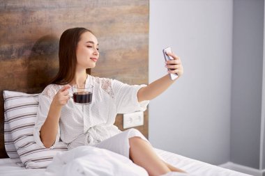 Güzel bir kadın yatakta bir fincan kahveyle selfie çeker.