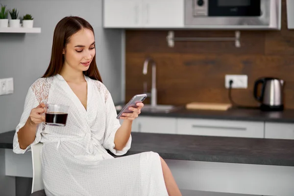 Lachende dame kijkt naar scherm van smartphone en drinkt koffie — Stockfoto