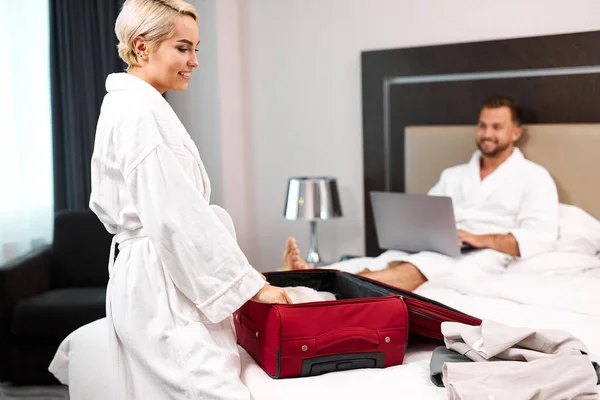 Young lady packing the suitcase while husband is working on laptop — Stock Photo, Image