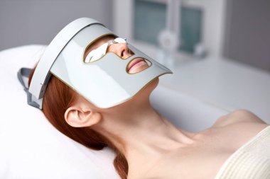 LED maskeli kadın estetik kliniğinde cilt gençleştirme terapisi görüyor.