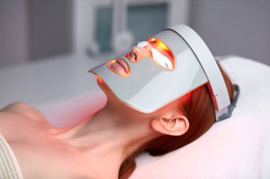 LED maskeli kadın estetik kliniğinde cilt gençleştirme terapisi görüyor.