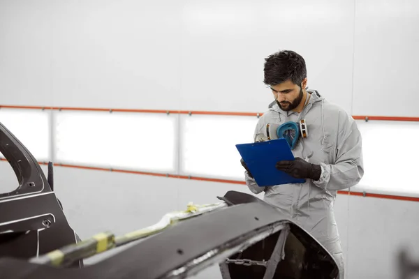 Knappe jonge Kaukasische auto monteur in uniform is het maken van notities tijdens het onderzoek van de auto in de auto service — Stockfoto