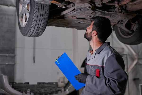 Handsome young caucasian auto mechanic in uniform is making notes while examining car in auto service — Stock Photo, Image