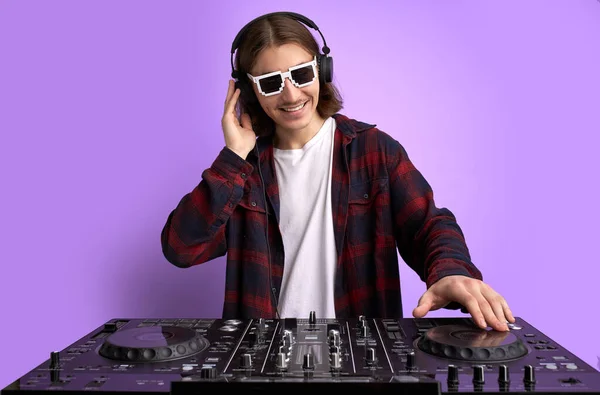 caucasian stylish dj man in sunglasses mixing console on purple studio background.