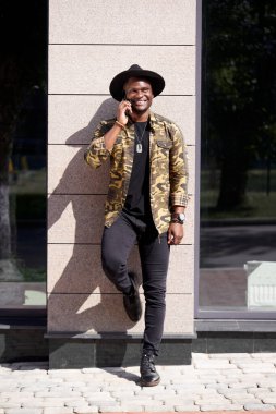 Esmer tenli siyah adam, günlük kıyafetlerini giyiyor, yürüyor ve telefonda konuşuyor.