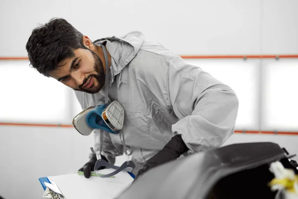 Knappe jonge Kaukasische auto monteur in uniform is het maken van notities tijdens het onderzoek van de auto in de auto service — Stockfoto