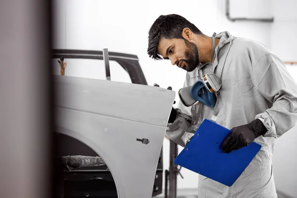 Knappe jonge Kaukasische auto monteur in uniform is het maken van notities tijdens het onderzoek van de auto in de auto service — Stockfoto