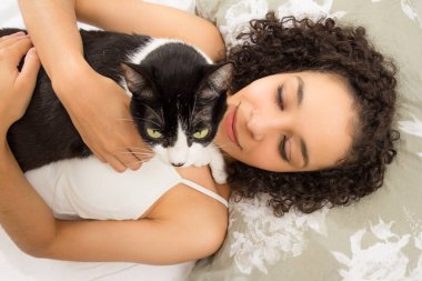 Yatakta yeşil gözlü sevimli kedi yavrusuyla eğlenen ve kucaklaşan siyah bir kadın. Hayvanlara sevgi, ilgi, sükunet, huzur kavramı