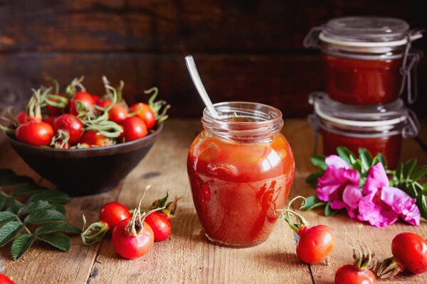 A jar of rose hip jelly and fresh rose hips on wooden table. 