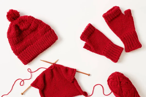 Knitting project in progress. Winter hat and mittens knitted with red yarn on white background.