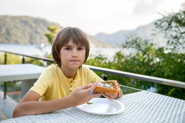 the boy eats waffles at a table in a street cafe by the sea