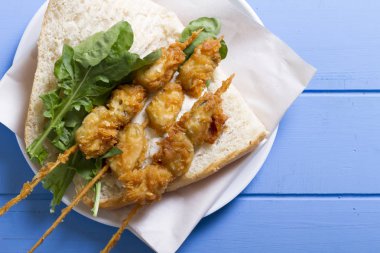 Lezzetli altın derin kızarmış pirzola midye şişleri yoğurt sos tartar ile hizmet veren sandviç, domates, dilimlenmiş beyaz soğan, maydanoz ve yeşil roka salatası mavi rustik masa arka plan üzerinde. Midye tava ekmek.