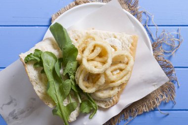 Lezzetli derin kızarmış halkalar sandviç hizmet beyaz daire plaka, yarım ekmek, soğan ve yeşil roka salatası rustik mavi ahşap arka plan üzerine dilimlenmiş. Kalamar çok mutfaklarının yenir. Üstten görünüm.