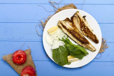 Lezzetli ızgara fileto levrek porsiyon dilimlenmiş soğan, domates, yeşil roka salatası ve sulu limon rustik mavi ahşap masa arka plan üzerinde oval beyaz plakalı füme. Alan, kopyalama kimse.