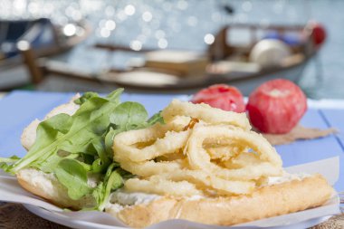 Lezzetli derin kızarmış halkalar sandviç hizmet beyaz daire plaka, yarım ekmek, soğan ve yeşil roka salatası rustik mavi ahşap arka plan üzerine dilimlenmiş. Kalamar çok mutfaklarının yenir. Üstten görünüm.