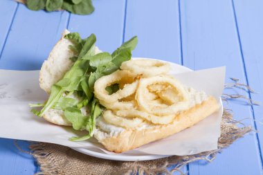 Lezzetli derin kızarmış halkalar sandviç hizmet beyaz daire plaka, yarım ekmek, soğan ve yeşil roka salatası rustik mavi ahşap arka plan üzerine dilimlenmiş. Kalamar çok mutfaklarının yenir. Üstten görünüm.